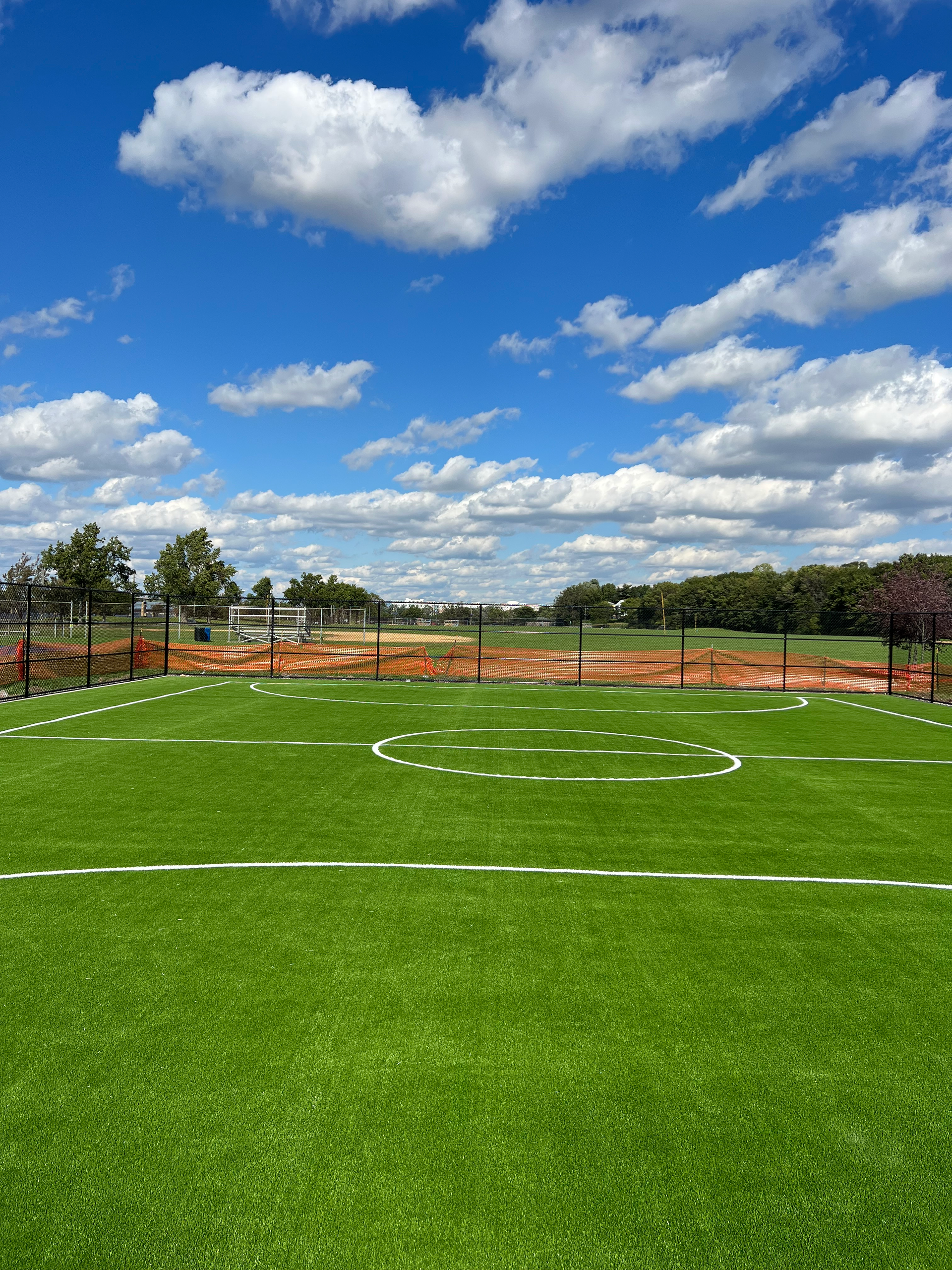 A grassy field with white painted soccer markings under a vibrant blue sky with scattered clouds and construction behind.