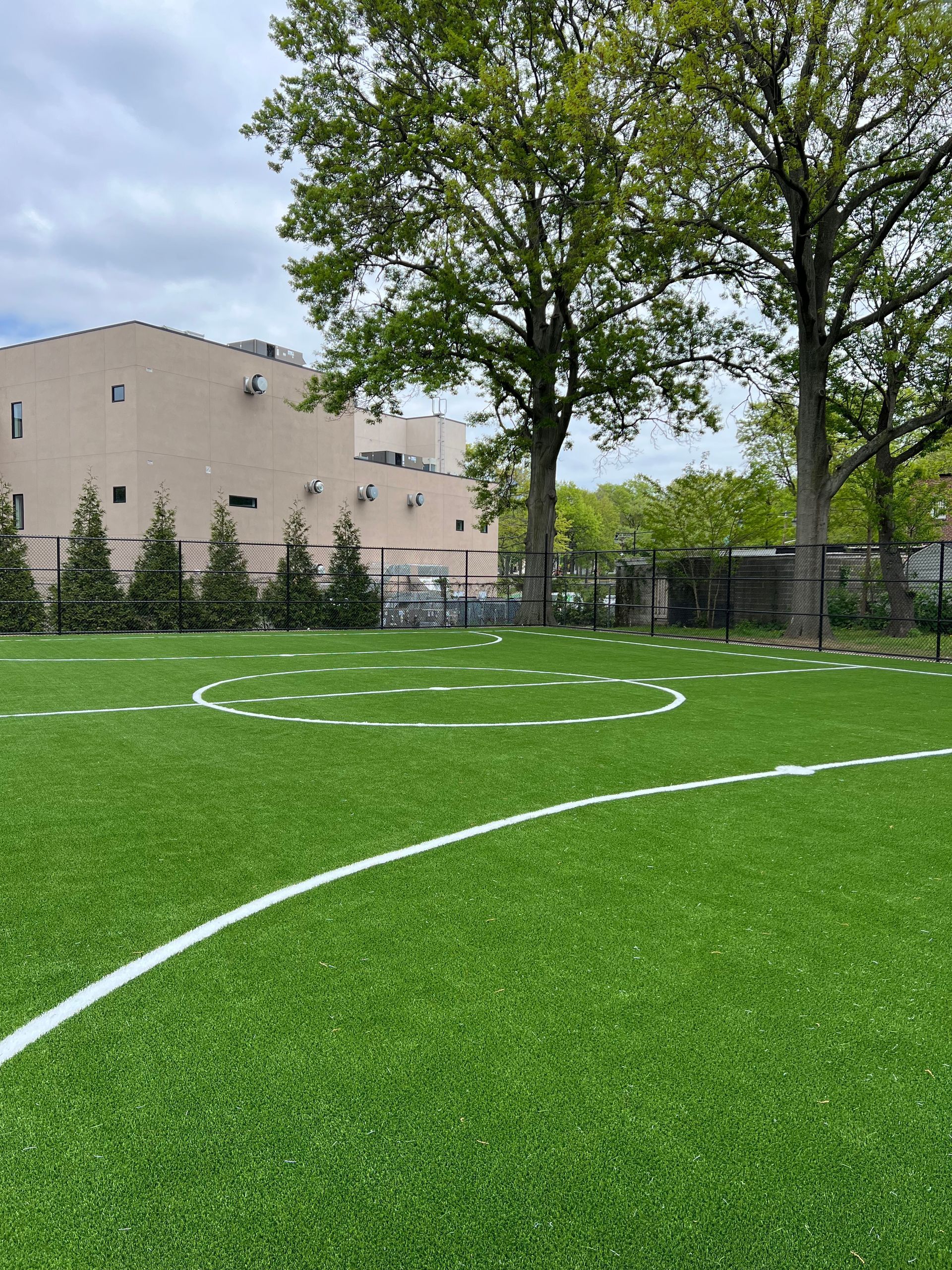 Artificial turf field with white line markings, surrounded by a metal fence and tall trees under a cloudy sky.