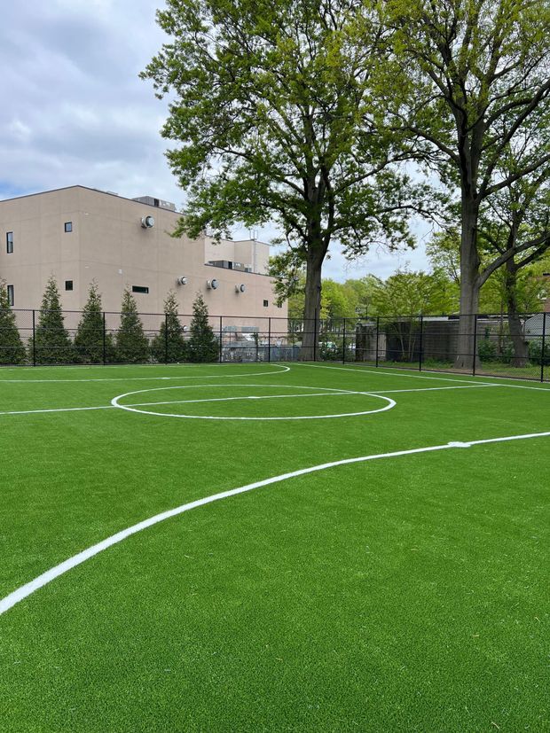 Artificial turf athletic field with white markings, surrounded by a metal fence and trees, with a building in the background.