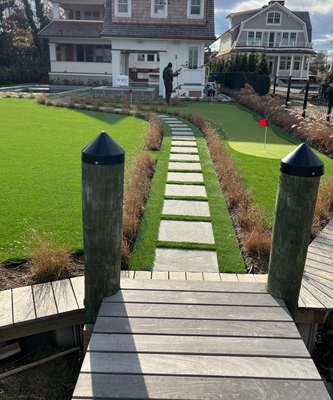 A stone stepping-stone path leads from a wooden dock across a residential lawn toward a house with a small putting green.