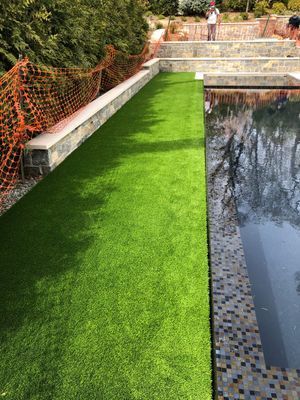 Artificial turf pathway alongside a stone-tiled swimming pool and a tiered stone wall under a protective orange mesh fence.
