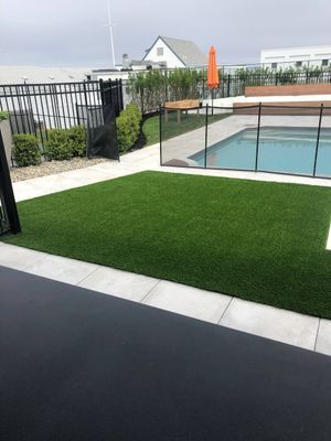 A rectangular patch of green artificial turf borders a pool area with a black safety fence and a bright orange umbrella.