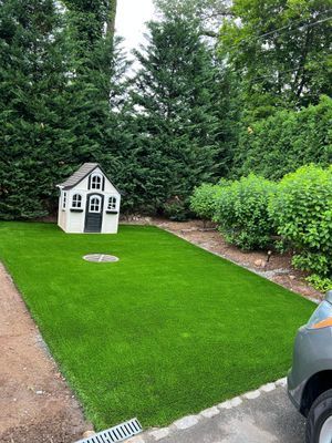 A white playhouse stands on a lush green artificial lawn next to a hedge, with a paved area and drain in the foreground.