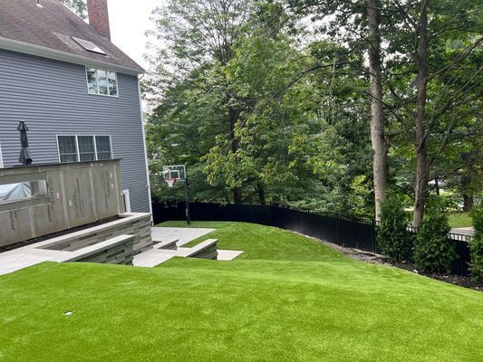 A lush green backyard with artificial turf, a gray house, stone patio, fire feature, and trees behind a black fence.