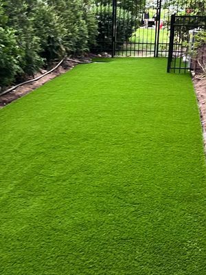 A narrow strip of vibrant green artificial turf lawn bordered by a tall hedge and a black metal fence leading to a gate.