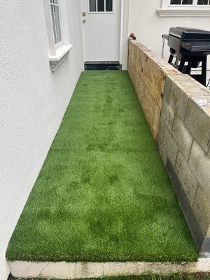 A narrow walkway surfaced with artificial grass leading to a white door, bordered by a house wall and a concrete block wall.