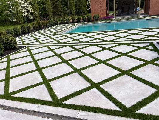 A patio featuring square concrete pavers arranged in a diamond pattern, separated by strips of lush green artificial grass.