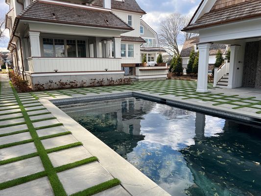 A swimming pool sits in a backyard next to a house, surrounded by square concrete pavers with grass growing in between.