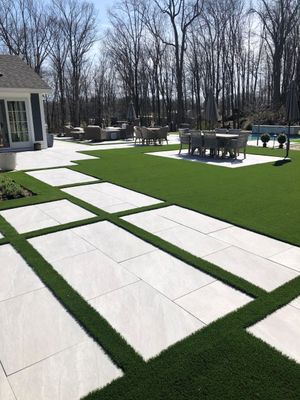 A modern backyard patio with rectangular light-gray stone pavers set in artificial grass, featuring a dining area.