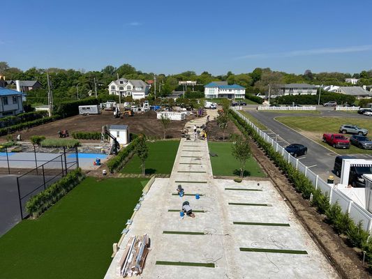 An aerial view shows workers laying turf sections on a long, concrete path in a landscape construction project.