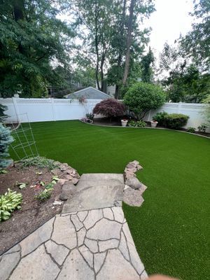 A manicured green lawn in a fenced backyard with a stone path in the foreground and a few trees in the distance.