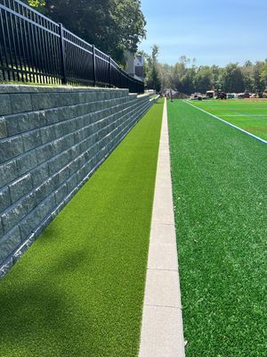 A stone retaining wall with a black fence runs along an artificial turf field, separated by a straight concrete pathway.