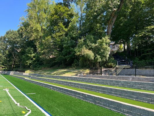 Artificial turf field with concrete tiered seating and stairs leading up a grassy hill lined with trees under a blue sky.