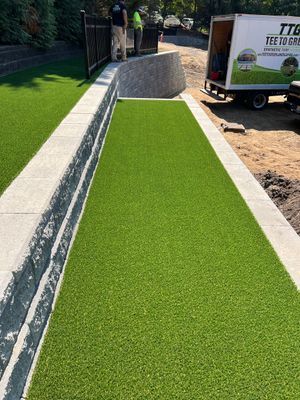 A newly installed strip of artificial turf bordered by a stone wall, with workers standing in the background near a truck.