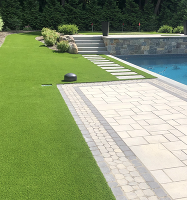 A paved pool deck beside a green lawn with stone steps leading to a raised garden area and a blue swimming pool.