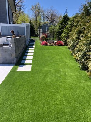 A backyard featuring a square-tiled path, a green artificial lawn, and a small garden bed with red flowers near a fence.
