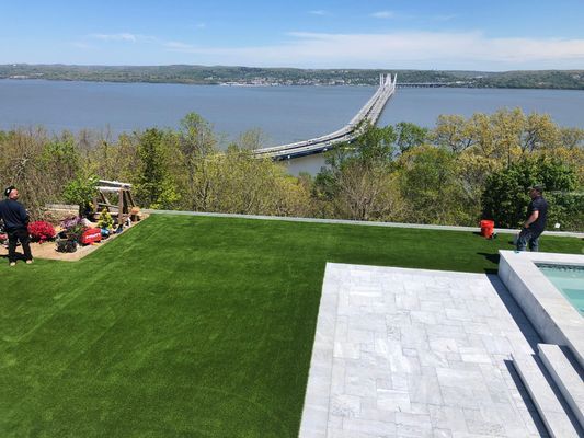 A patio with a green lawn overlooks a large body of water and a long bridge spanning across it on a sunny day.