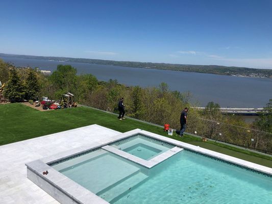 An infinity pool and spa overlook a large river and bridge on a sunny day, with two people working on the lawn nearby.