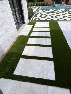 A path of rectangular white stone pavers set in artificial green turf alongside a modern house and pool area.