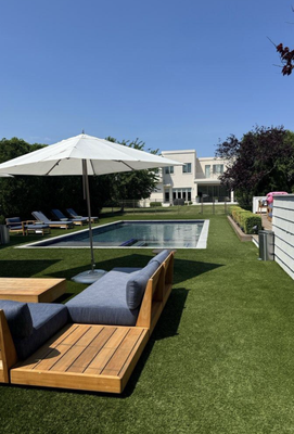 A swimming pool set in a sunny backyard with lounge seating, a white umbrella, and a large house in the background.