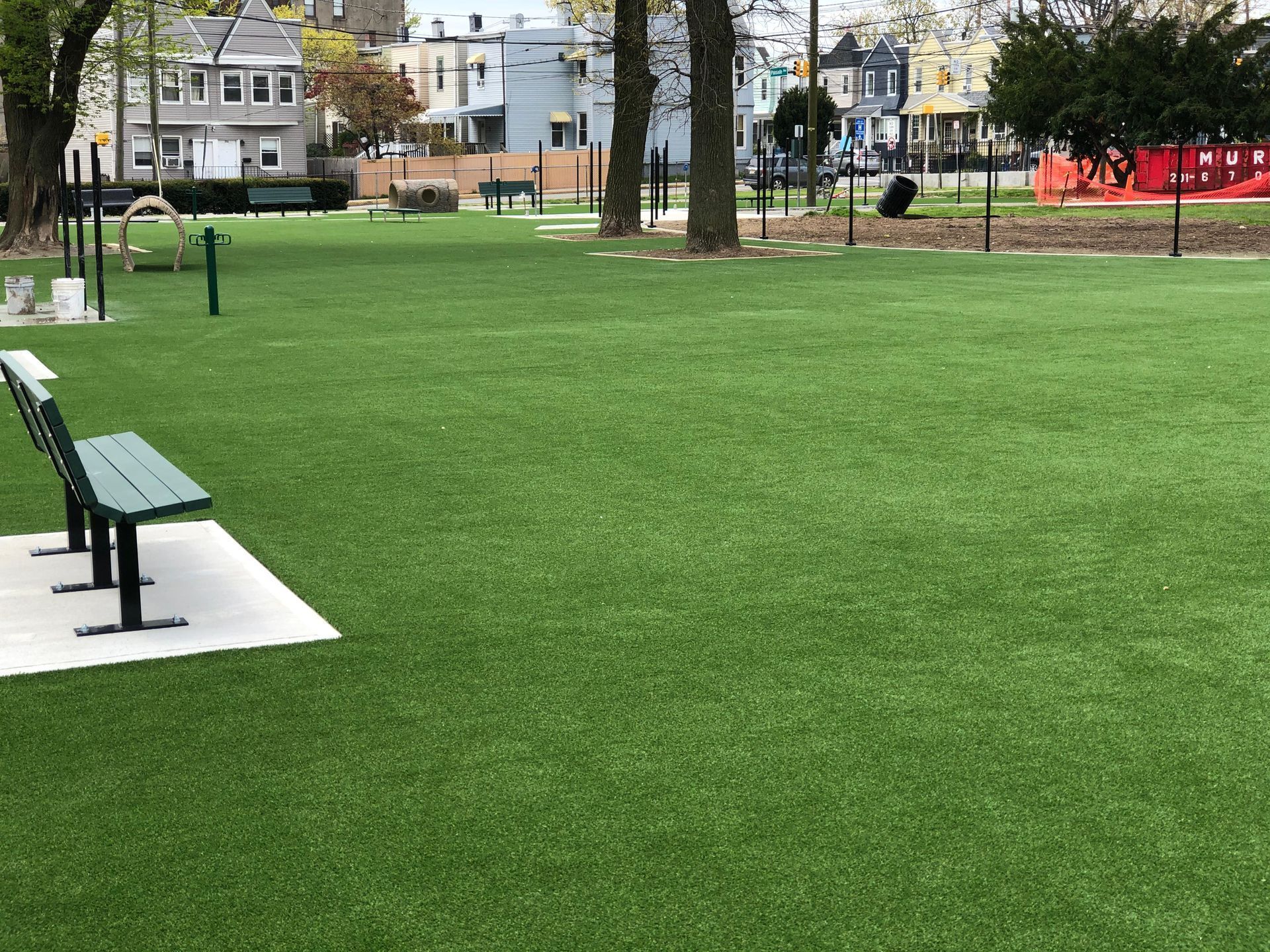 A green bench sits on a concrete pad in a public park with artificial grass, surrounded by trees and residential houses.