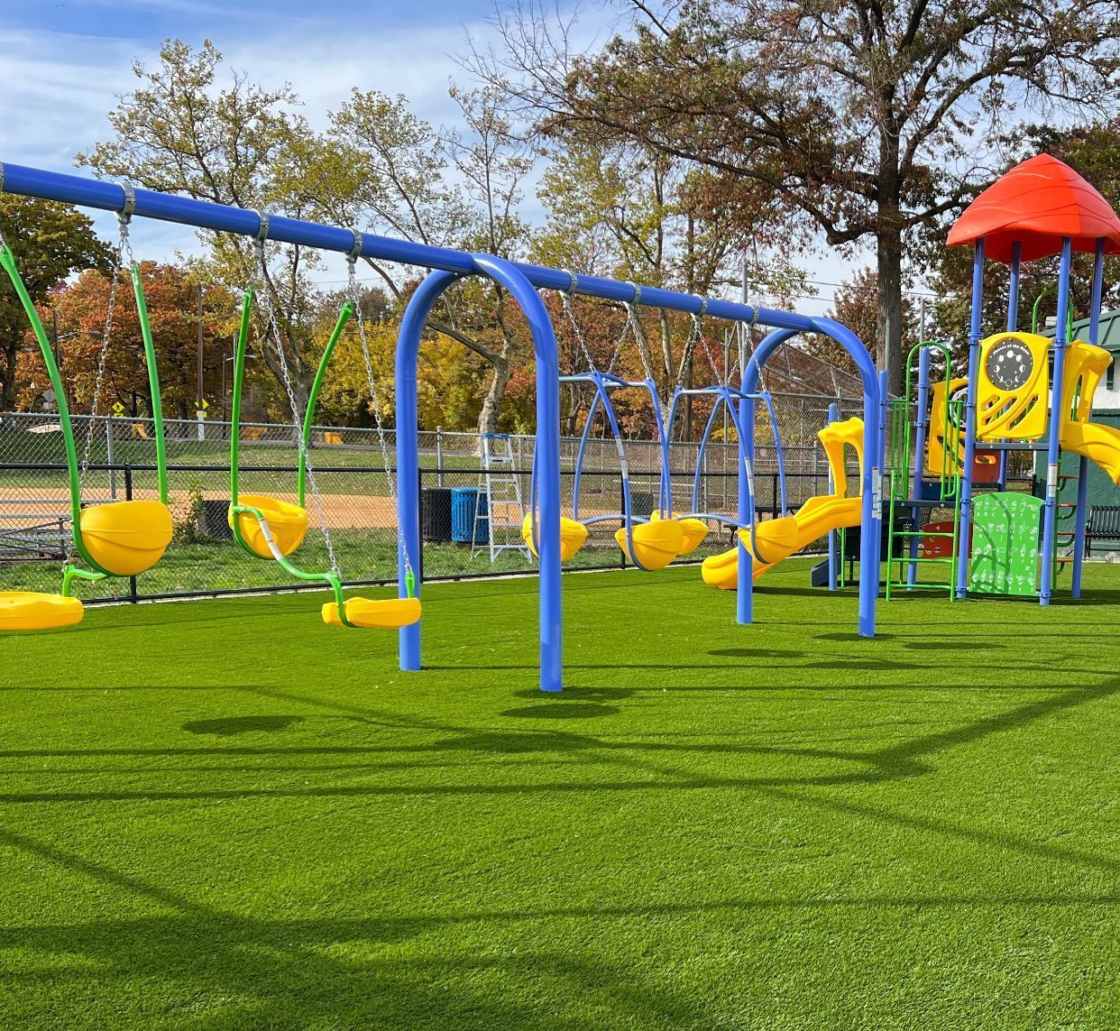 Playground with yellow bucket swings hanging from a blue frame, with a jungle gym in the background on green turf.