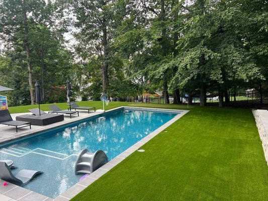 A rectangular swimming pool surrounded by a manicured green lawn and tall trees, with lounge chairs on the patio.