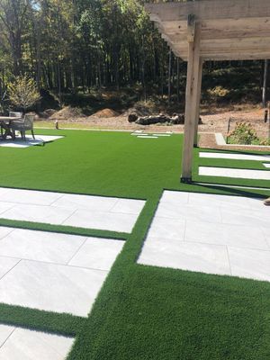 A backyard with light-colored paver stones set into a vibrant green artificial lawn beneath a wooden pergola.