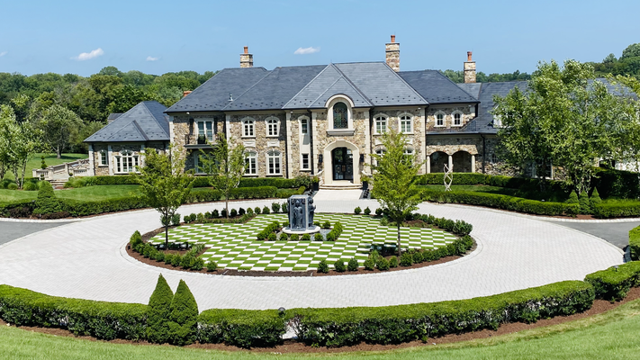 A large, multi-story stone mansion with a slate roof and a circular driveway featuring a central garden sculpture.