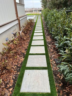 A stone paver walkway with grass inlays, bordered by a building, fallen leaves, and a hedge, leading toward a pool area.