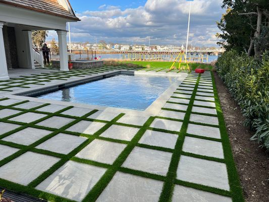 A rectangular swimming pool surrounded by a patio of square stone pavers with green grass growing between them.
