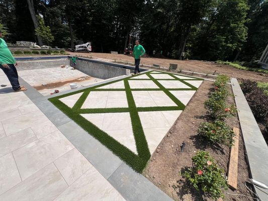 Two people stand near a swimming pool construction site featuring a geometric patio design with grass-filled seams.