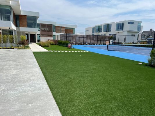 Modern buildings with large windows overlook a green lawn and a bright blue tennis court under a light blue sky.