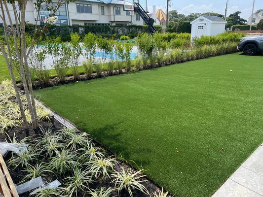 A lush green artificial lawn bordering a garden bed with variegated plants and a hedge, with a small white shed in back.