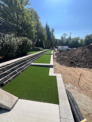 Newly installed artificial turf sections bordered by a concrete retaining wall, next to a dirt mound under a clear blue sky.
