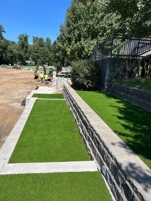 Workers construct an artificial turf terrace with stone retaining walls next to a dirt construction site.