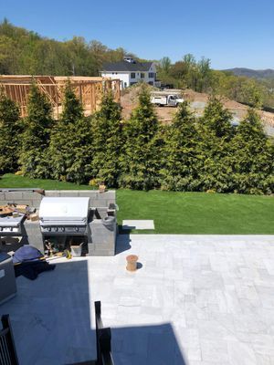 A stone patio with an outdoor kitchen overlooks a green lawn, a row of tall evergreen trees, and a house under construction.