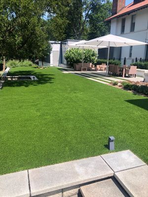A wide green lawn in front of a white house with an outdoor patio area featuring tables, chairs, and umbrellas.