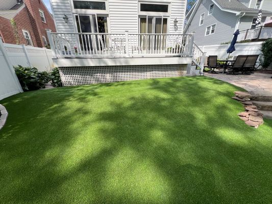 A lush green artificial lawn in a backyard, situated below a white deck with a staircase leading to a patio area.