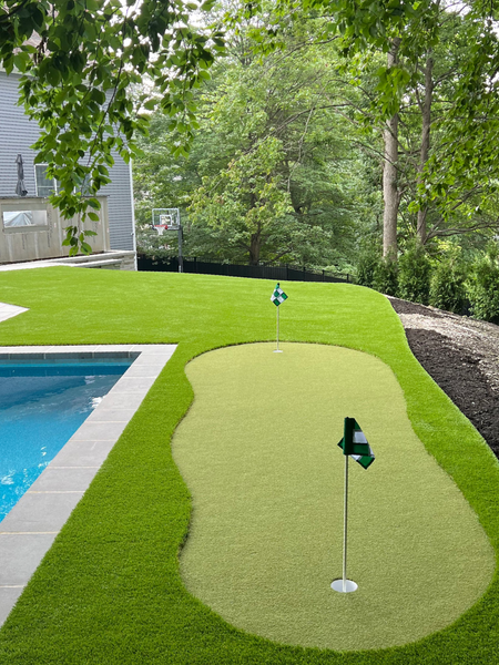 A backyard putting green with two holes and flags next to a swimming pool, surrounded by manicured grass and trees.