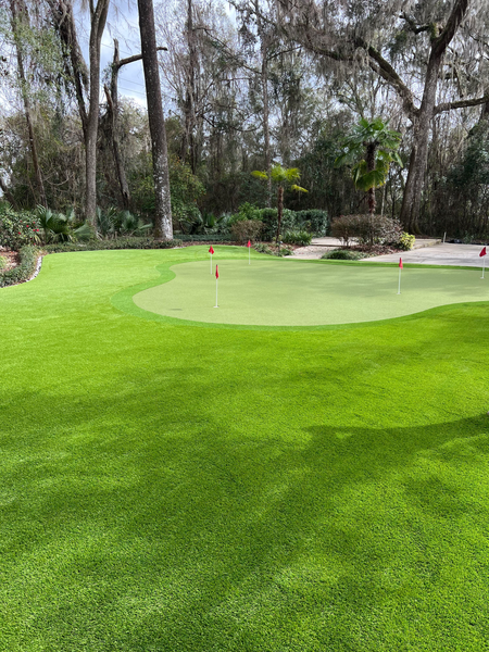 A vibrant green artificial putting green with multiple red golf flags, set in a backyard surrounded by trees.