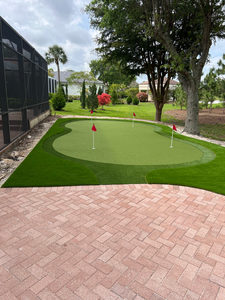 A backyard putting green with red flags, artificial grass, and surrounding landscaping, viewed from a brick patio.