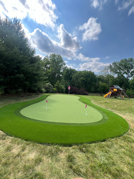 A backyard putting green with multiple holes and flags, surrounded by grass, trees, and a wooden playset under a blue sky.
