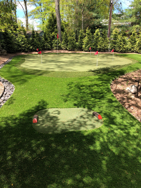 A backyard putting green with a tee box, several golf holes with red flags, and surrounding trees.