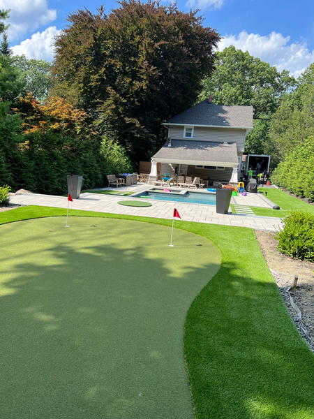 A backyard putting green with two red flags, leading to a stone patio with a swimming pool and a two-story house.