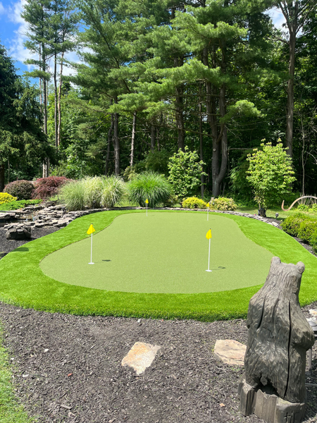 A backyard putting green with two yellow flags, surrounded by landscaping, trees, and a wooden bear carving.