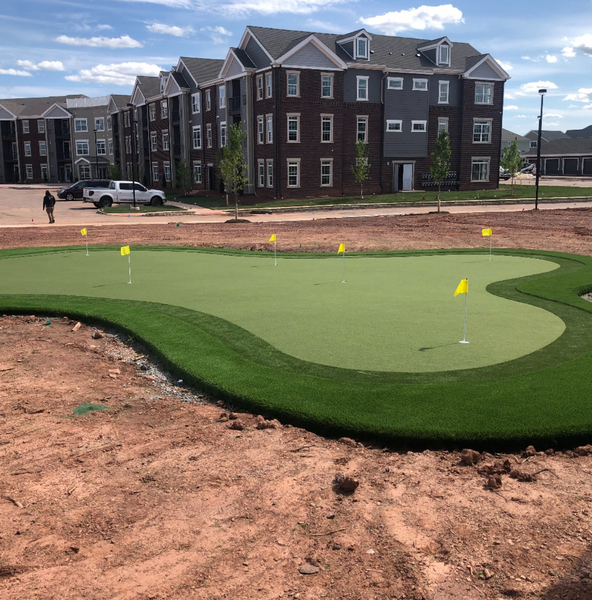 A putting green with yellow flags sits on a construction site in front of a modern three-story brick apartment building.