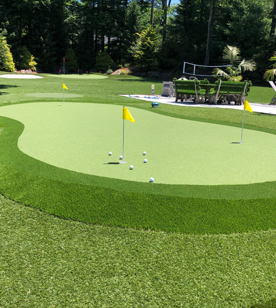 A backyard putting green with several golf balls on the turf, two yellow flags, and outdoor seating in the background.