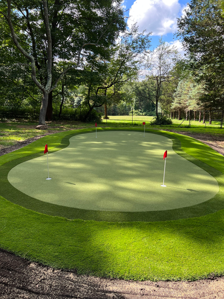 A synthetic putting green with three red flags, surrounded by lawn and trees under a blue sky with white clouds.