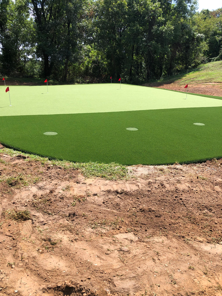 A backyard putting green with light and dark green artificial turf, red flags, and surrounding unfinished landscaping.